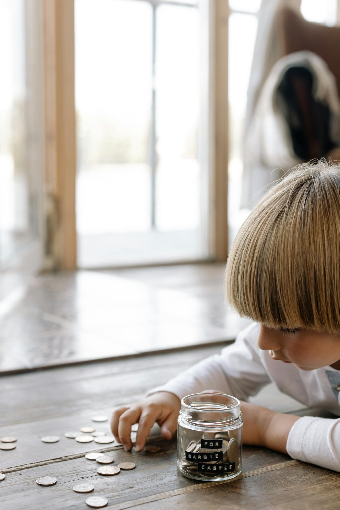 A young boy counting coins and placing them into a jar labeled 'for barbie castle' indoors on wooden flooring.